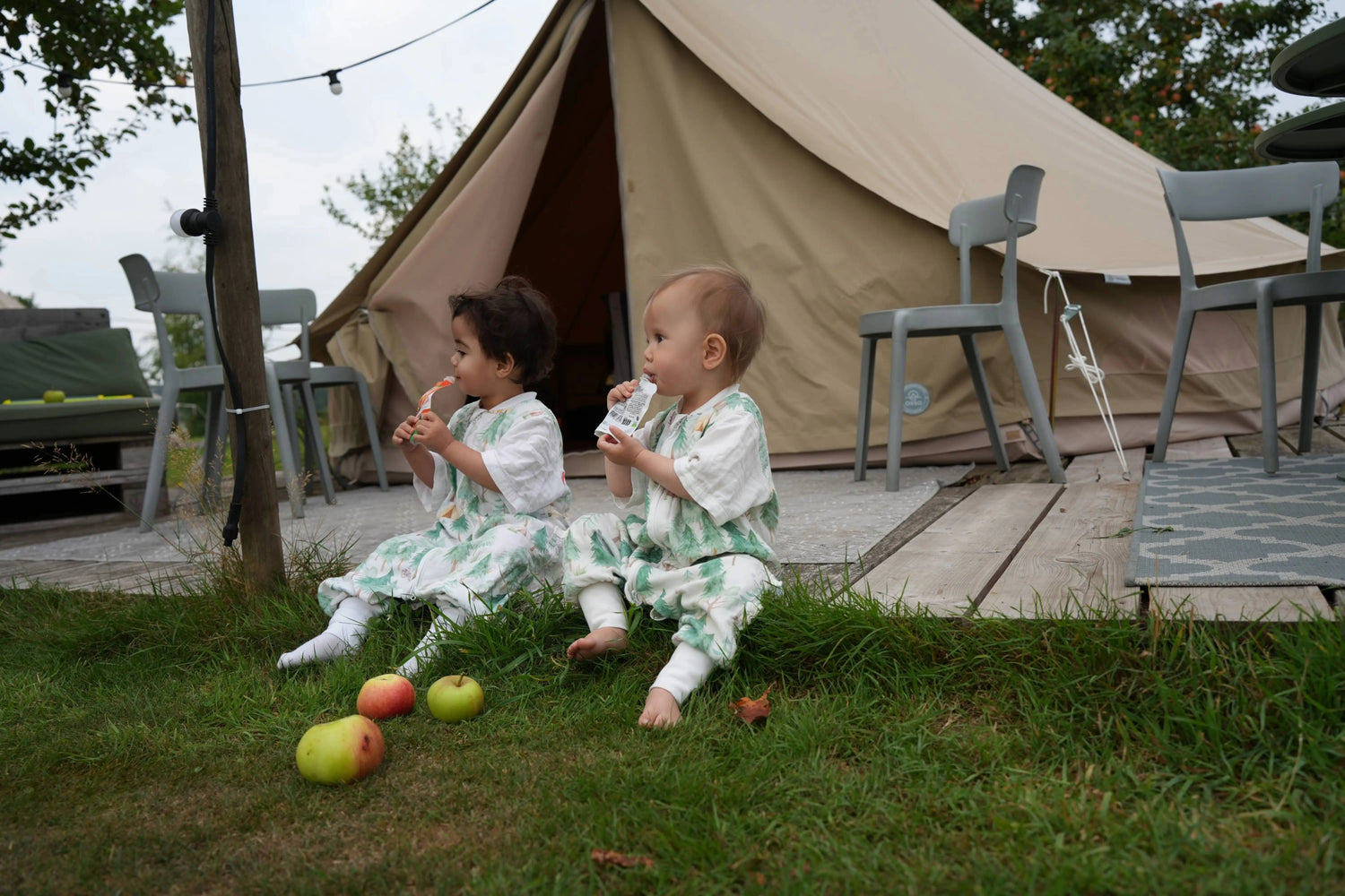 Two children in vosbos sleep suit sitting on grass in front of a tent with apples around them.